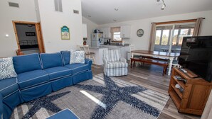 A living room with a blue sofa, a star-patterned rug, a wooden TV stand, and a kitchen in the background. - Oceanside, Private Pool, Sun Decks, 660 ft to beach access! -WW951, Momma's Happy, Daddys Broke (Corolla)