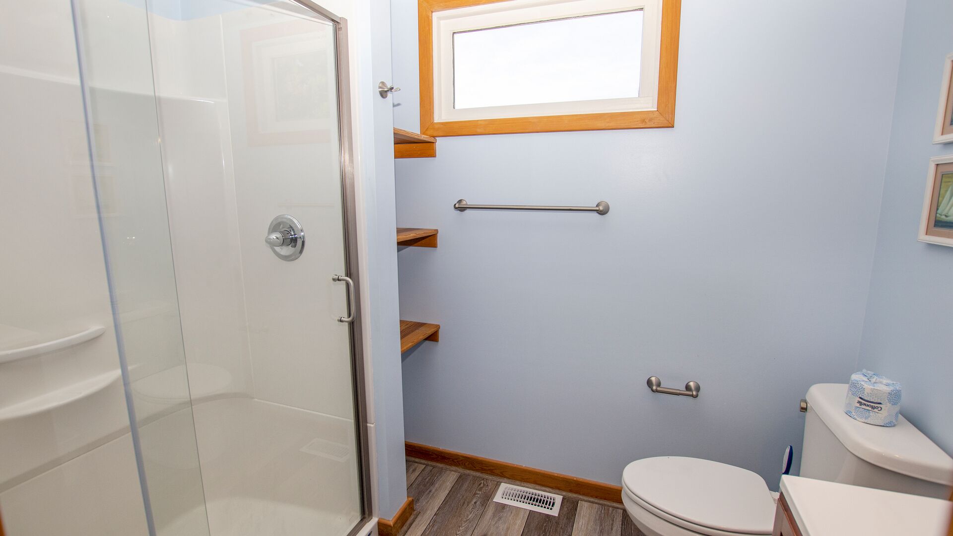 Une salle de bain avec une douche à parois de verre, des toilettes avec un porte-rouleau de papier toilette et une fenêtre au-dessus du lavabo.
