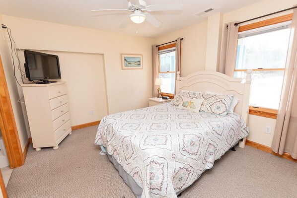 A bedroom with a bed, a TV on a dresser, a ceiling fan, a window with curtains, and a wall-mounted picture.