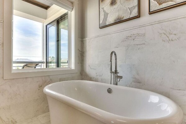 Un baño con una bañera independiente, un ventanal con vistas y dos cuadros abstractos en la pared.