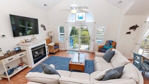 A spacious living room with a large sofa, a fireplace, a television, and a balcony with outdoor seating. - Oceanside, Priv Pool, Hot Tub, OS Shuffleboard, Rec Rm, WW1039, Island So Remote (Corolla)