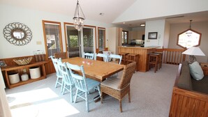 A spacious dining area with a wooden table and light blue chairs, a kitchen area with wooden cabinets, and a seating area with a sofa and cushions. - Oceanside, Private Pool, Hot Tub, Screened Porch, 650 ft to Beach Access, WW1021, Zip-A-Dee-Dune-Dah (Corolla)