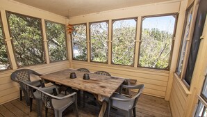 A wooden deck with a table and chairs, large windows, and a view of trees. - Oceanside, Community Amenities, Hot Tub, Sun Decks, 117, Sound Waves (Corolla)
