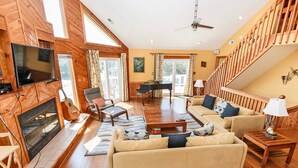 A spacious living room with a fireplace, a television mounted on a wooden wall, a piano, and a staircase leading to an upper level. - Oceanside, Community Amenities, Hot Tub, Sun Decks, 117, Sound Waves (Corolla)