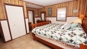 Un dormitorio con piso de madera, una cama con colcha estampada, cabecera de madera, mesita de noche, cómoda, espejo y ventana con persianas.