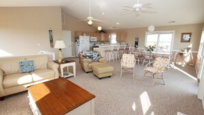 A spacious living room with a sofa, ottoman, and coffee table, a kitchen area with wooden cabinets, and a dining area with multiple chairs.