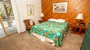 A bedroom with a wooden sloped ceiling, a bed with floral bedding, two bedside lamps, a wicker chair, and a green dresser. - Oceanside, Priv Pool, H Tub, Close to Beach Access, OS56H, Slapwater (Corolla)