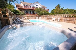 Ein Whirlpool mit einer Holzterrasse und Liegestühlen, umgeben von einem Holenzaun, mit einem Haus im Hintergrund.