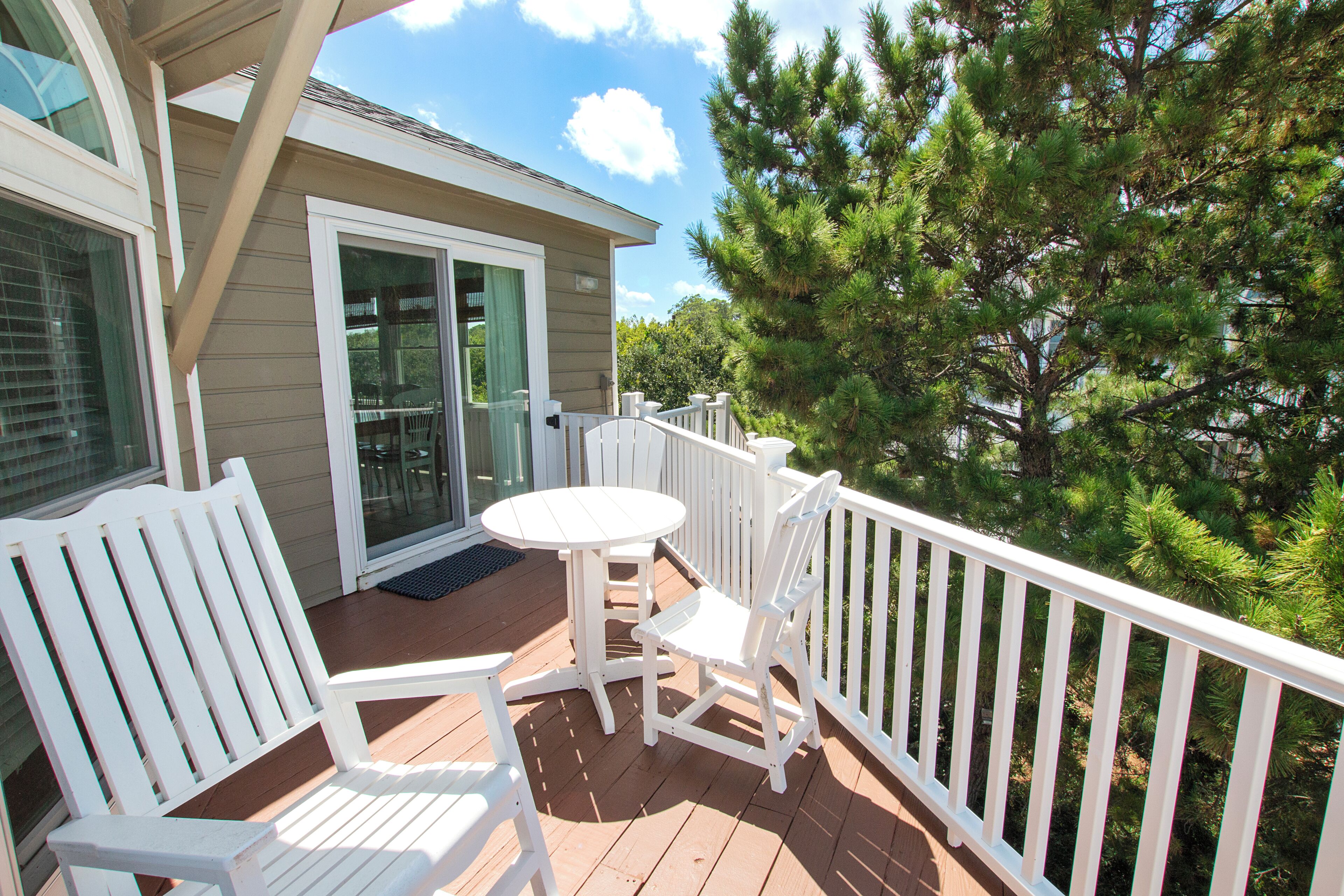 A wooden deck with a white table and chairs, a glass door entrance, and a view of pine trees.