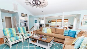 A spacious living room with a large brown sofa, two white armchairs with blue and yellow patterned cushions, a wooden coffee table, and a patterned rug.