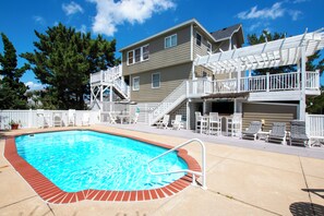 A swimming pool with a brick edge, surrounded by a deck with lounge chairs and a covered area with stairs leading to a building. - Oceanside, Private Pool, Hot Tub, Close to beach access, 224, Its All Good (Corolla)