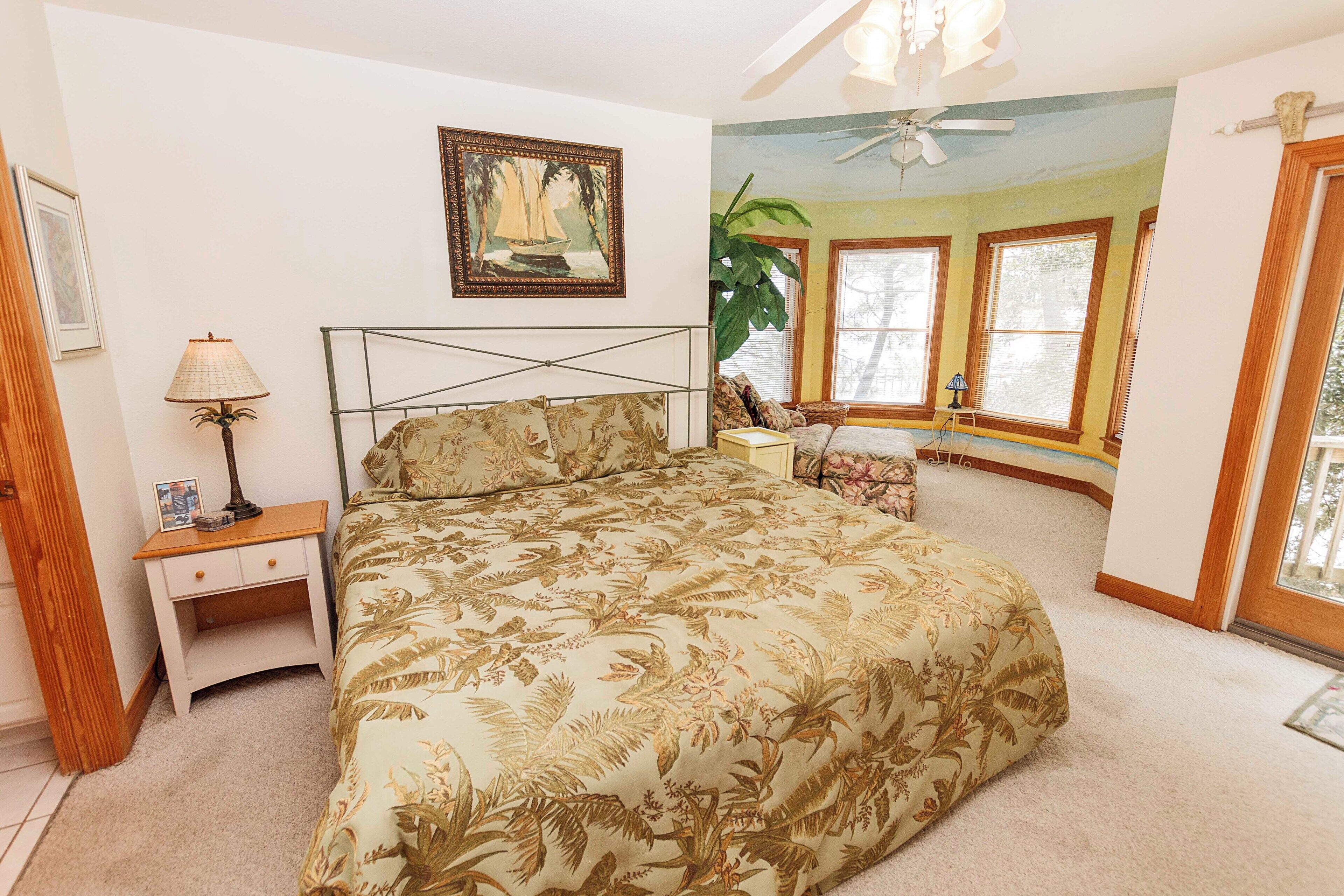 A bedroom with a bed, a nightstand, a lamp, a painting, and a large window.