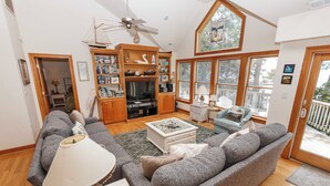 A spacious living room with a large gray sofa, a coffee table, and a television. There is a wooden cabinet with various items and a ceiling fan. - Soundfront, Sound views! Dogs Welcome, Private Pool! 520, Bay Spray (Corolla)