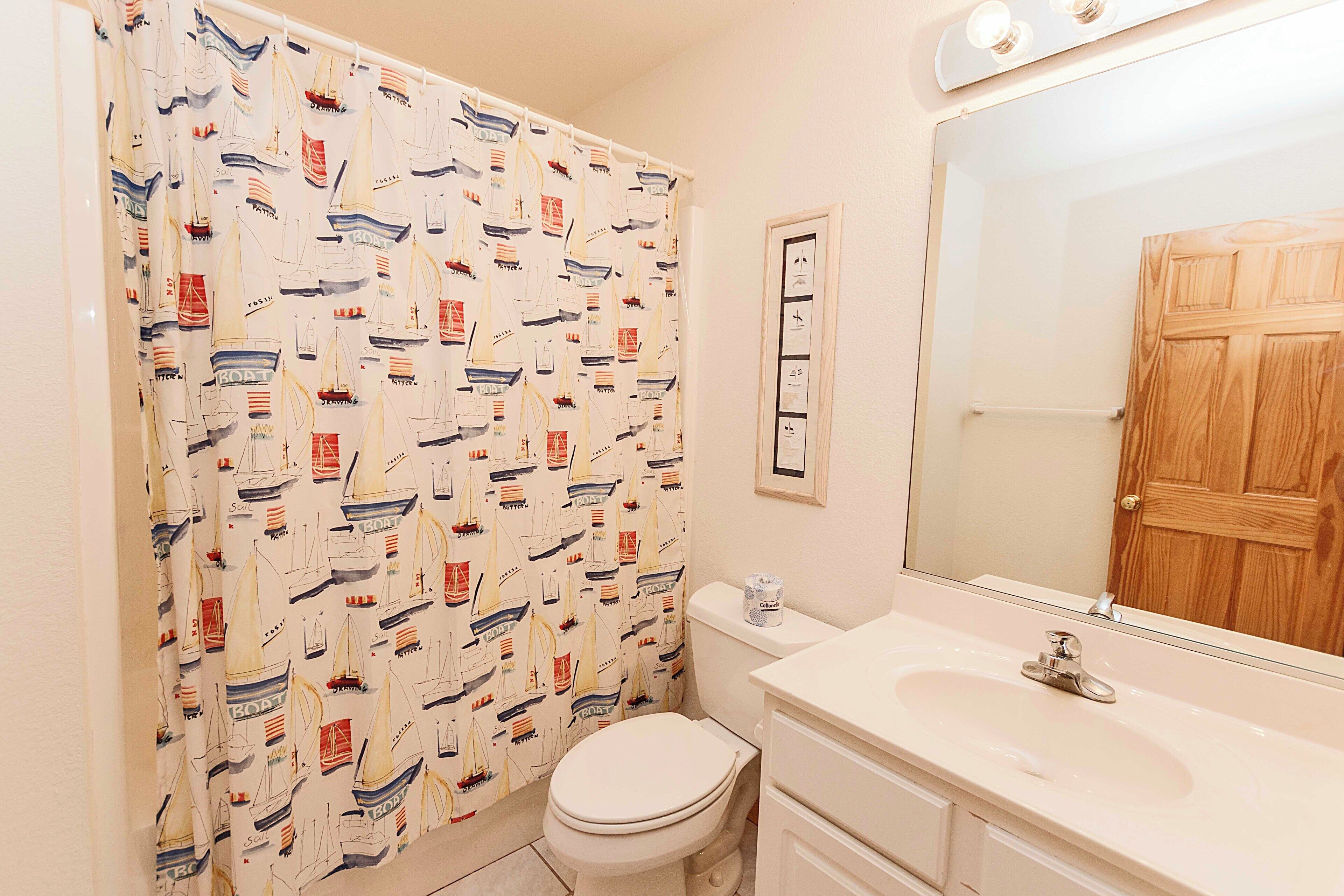 Un baño con cortina de ducha de temática náutica, inodoro blanco, lavamanos con espejo y puerta de madera.