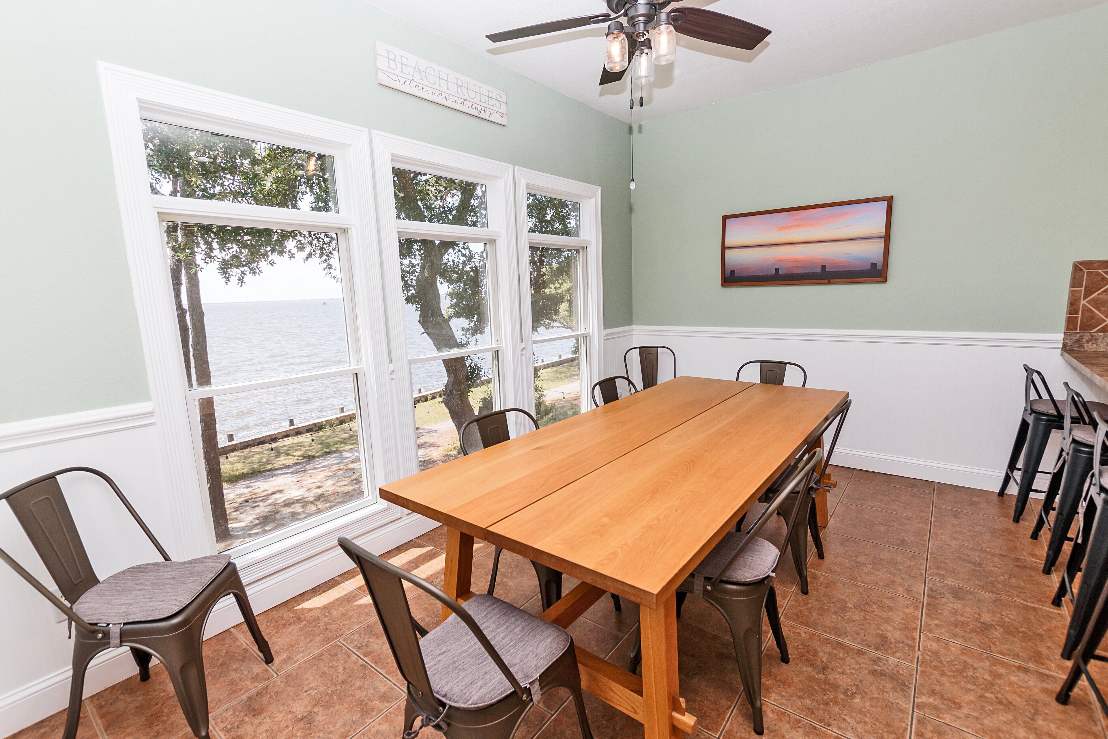 Un coin repas avec une table et des chaises en bois, de grandes fenêtres donnant sur la mer et un ventilateur de plafond.