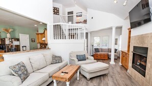 A spacious living area with a high ceiling, a staircase, a fireplace, and a kitchen area visible in the background. - Soundfront, Sound views! Private Pool, Community Amenities, 504, Lost N' Sound (Corolla)