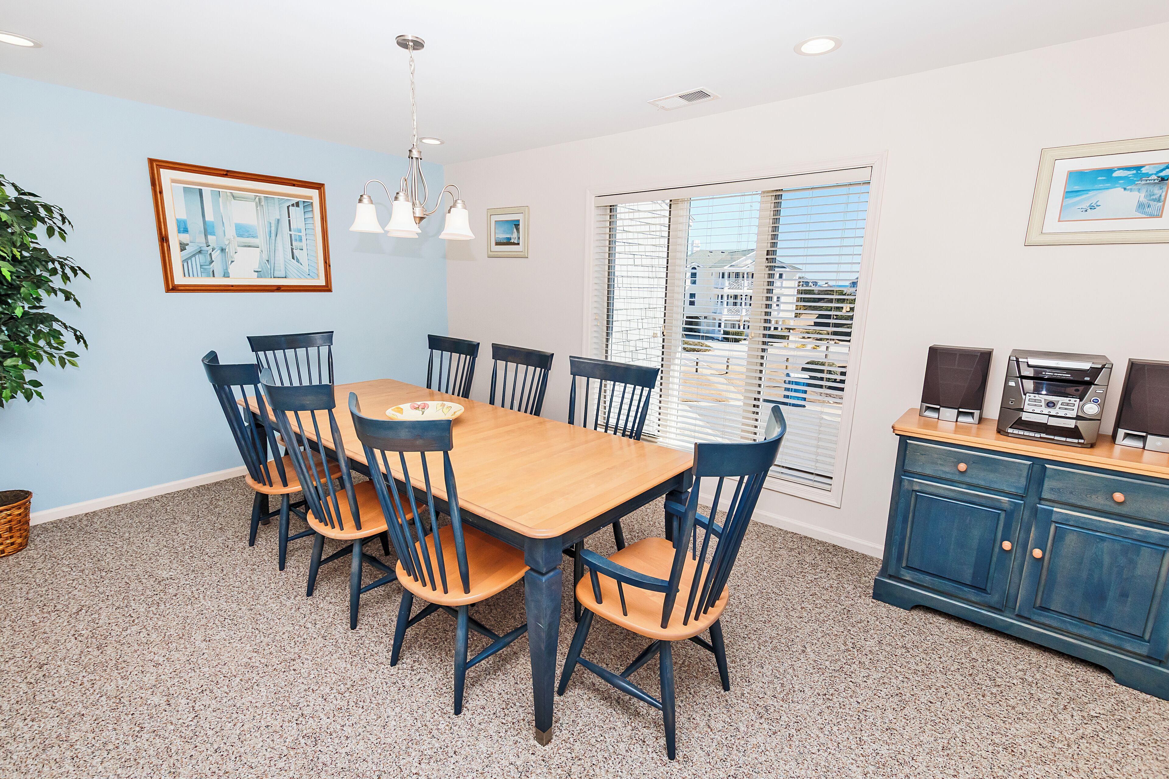 Un coin repas avec une table et des chaises en bois, un meuble bleu contenant des haut-parleurs et une fenêtre avec des stores.