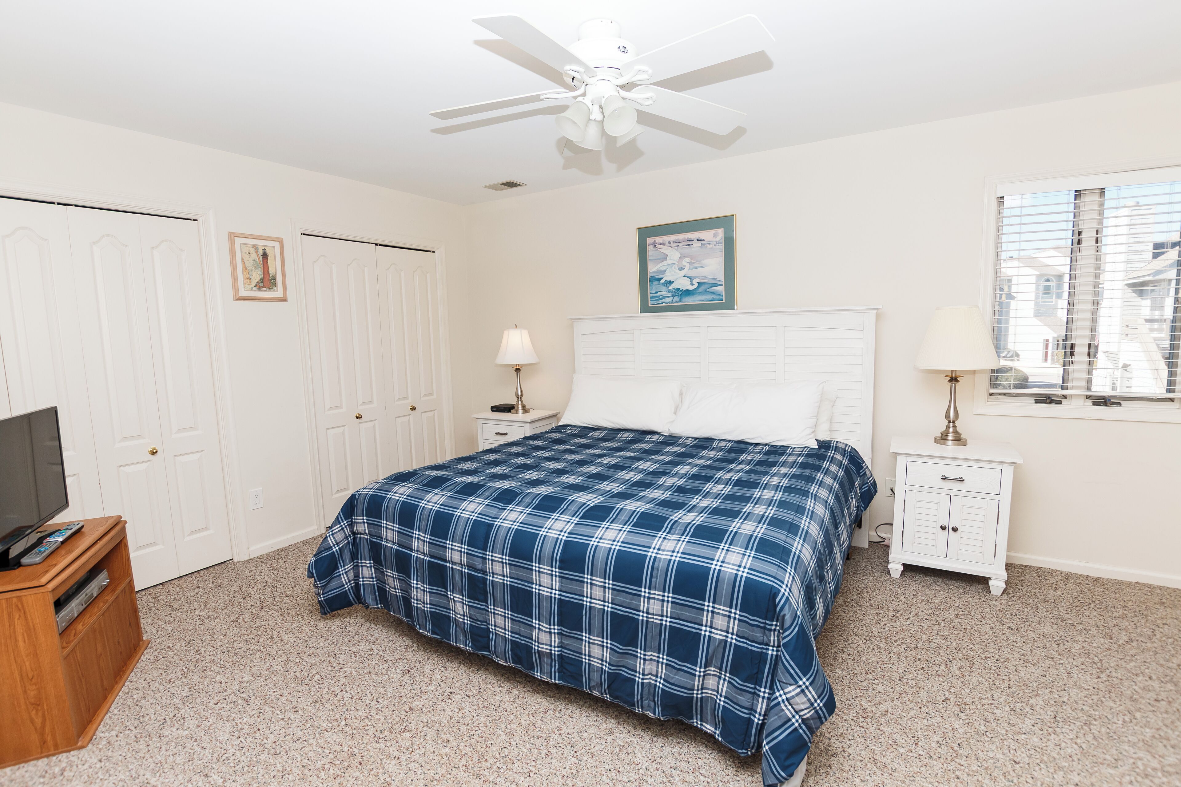 A bedroom with a bed, a ceiling fan, a nightstand, a TV stand, and a window with blinds.