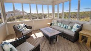 A covered patio with wicker furniture, a wooden coffee table, and a view of a house and trees. - Oceanside, Private Pool, Rec Room, Vllyball, PI157, Vintage Pine (Corolla)