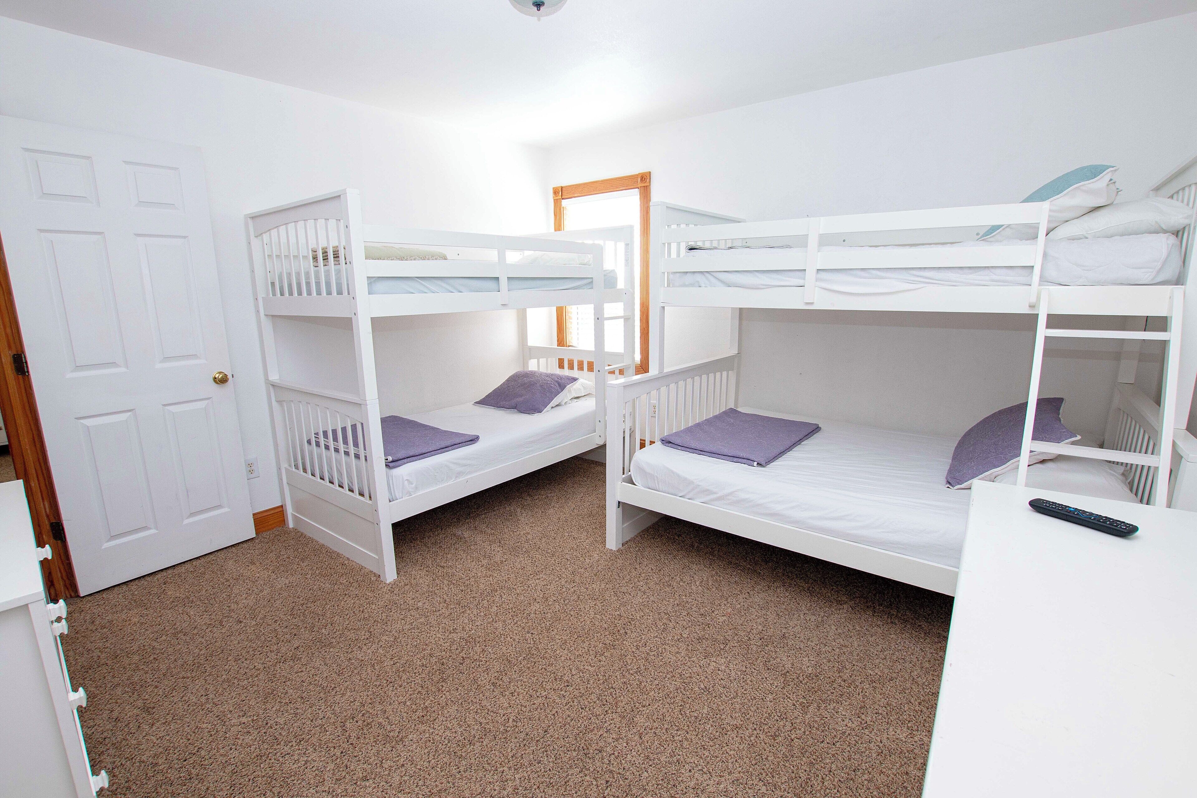 A room with two bunk beds, a white door, and a white desk with a remote control.