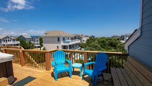 A wooden deck with two blue Adirondack chairs and a small table, overlooking a residential area with houses and a clear blue sky. - Oceanside, easy walk to Beach, Hot Tub, Dogs Welcome, OL4, Lakes Ocean View (Corolla)