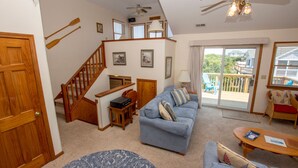 A cozy living room with a blue sofa, wooden staircase, and a view of the outdoors through a sliding glass door. - Oceanside, easy walk to Beach, Hot Tub, Dogs Welcome, OL4, Lakes Ocean View (Corolla)
