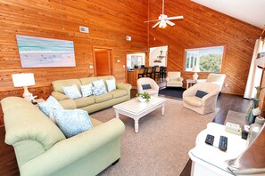 A spacious living room with a high ceiling, wooden walls, a large painting, and a dining area with chairs and a table. - Oceanside, 5 BRs, Pool, Hot Tub, Close to Beach Access, T10, Surf N Duck (Duck)