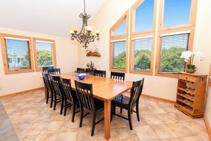 A dining room with a wooden table and chairs, a chandelier, and large windows with blinds.