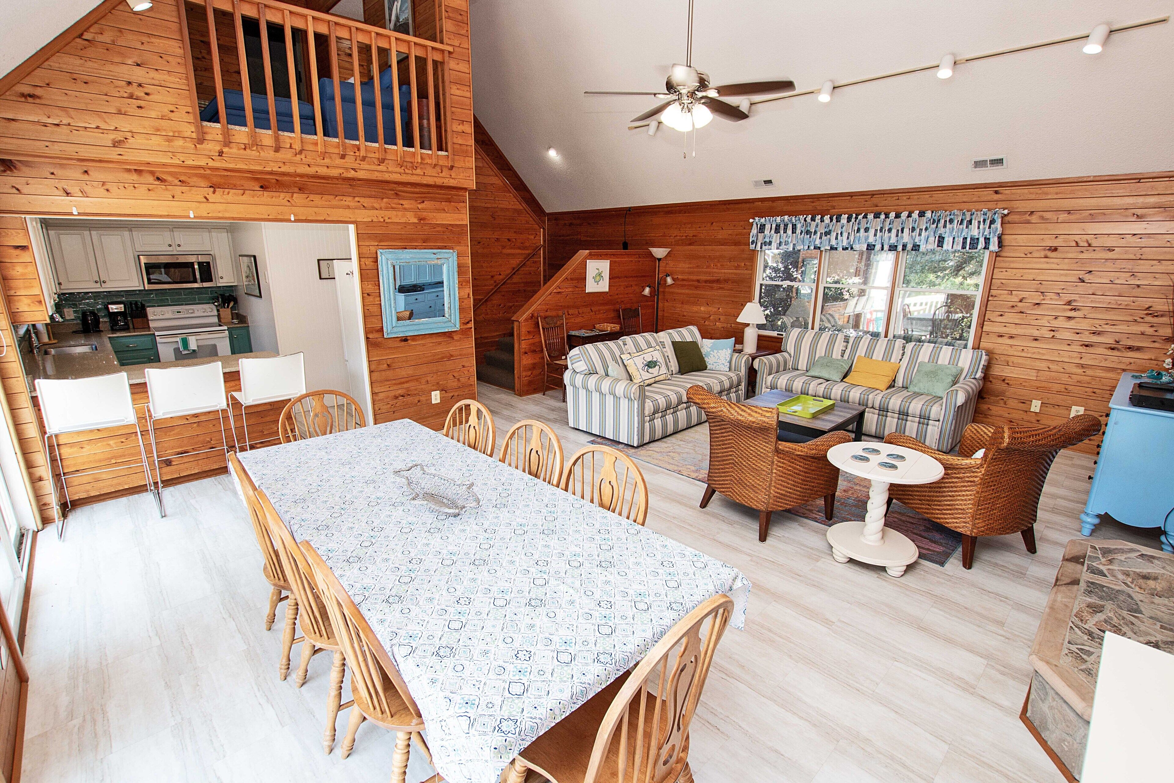 Amplia zona de estar tipo loft con mesa de comedor, sillas, sofá y una pequeña mesa redonda. Se ve una cocina al fondo con armarios de madera y un microondas.
