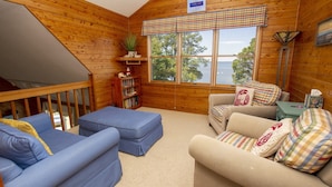 A cozy cabin interior with a wooden interior, a loft bedroom, a sofa, a chair, and a view of the outdoors.