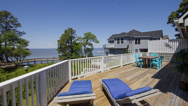 A wooden deck with two lounge chairs and a table, overlooking a body of water and a house.