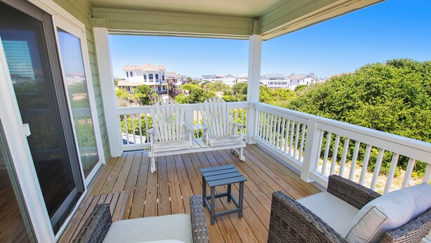A covered porch with a wooden deck, white railings, a bench, two wicker chairs, and a small table. - Oceanside, Pool, Hot Tub, 945 ft to Beach Access, WW935, Sea Watch (Corolla)