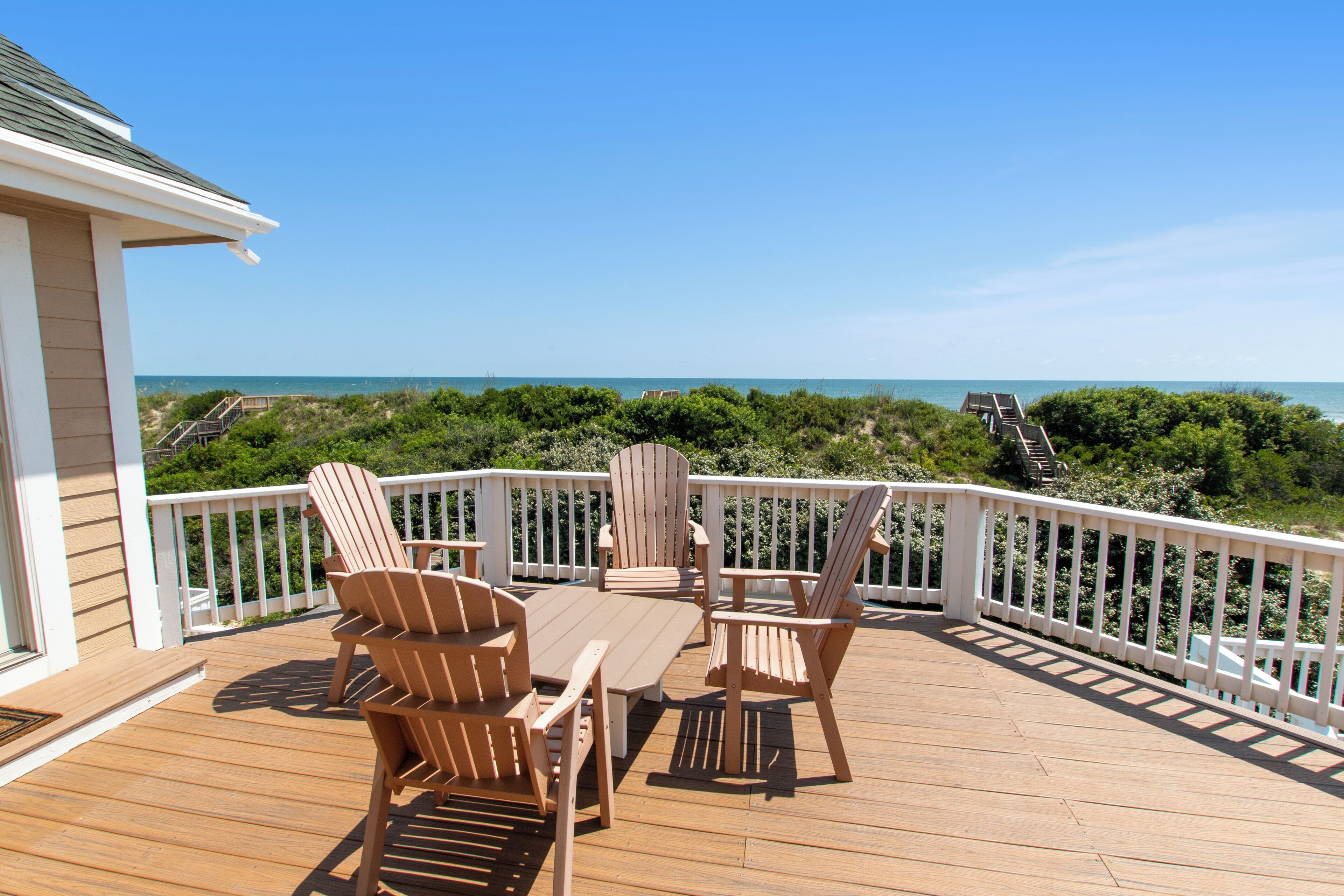 A wooden deck with a table and chairs, overlooking a beach with a clear view of the ocean.