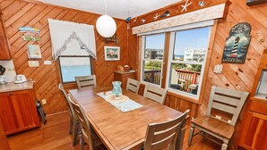 A wooden dining area with a table and chairs, a window with a view of buildings, and a sign indicating a raw bar and grill. - Oceanside, Close to beach, Sunroom, Carport, SNH351, Sea Sharp (Nags Head)