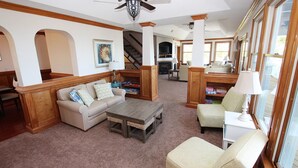A spacious living room with a beige sofa, a wooden coffee table, and a patterned armchair. There is a fireplace, a staircase, and a bookshelf filled with books. - Soundside, Priv Pool, H Tub, ELEV, Rec Rm, Theatr Rm, FR1, The Drake (Duck)