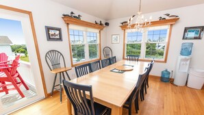 A dining room with a large wooden table, multiple chairs, and a chandelier. There are windows with views of greenery and buildings outside.