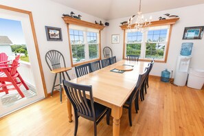 A dining room with a large wooden table, multiple chairs, and a chandelier. There are windows with views of greenery and buildings outside. - Semi-Oceanfront, 7 BRs, Priv Pool, H Tub, Rec Rm, WL1068, Footloose (Corolla)