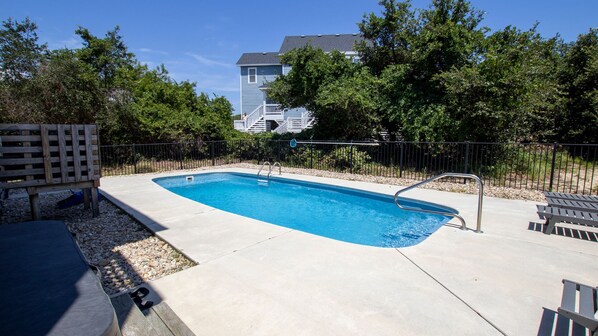 Une piscine rectangulaire avec un escalier en métal, entourée d’une terrasse en béton et d’une clôture en métal. On y trouve des bancs en bois et une chaise longue en bois près de la piscine.