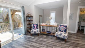 A spacious living room with a wooden floor, a glass door leading to a balcony, and a dining area with a wooden table and chairs. - Oceanside, 4 BRs, Private Pool, Hot Tub, Screened Porch, WC955, La Carolina (Corolla)