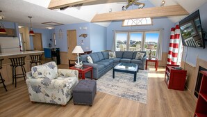 A spacious living room with a large sectional sofa, a matching armchair, and a coffee table. There is a kitchen area with a bar and a dining table. The room has a high ceiling with a fan and a large window offering a view of the outside.