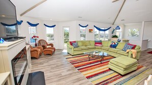 A spacious living room with a sectional sofa, a coffee table, and a TV. There are multiple windows with blue curtains, a fireplace, and a colorful striped rug. - Semi-Oceanfront, Rec Room, Private Pool, Ocean Views, WL1092, Maritime (Corolla)