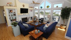 A spacious living room with a fireplace, large windows, and a view of the neighborhood. - Soundside, Pool, Hot Tub, Community Amenities, MS24, Turtle Whispers (Corolla)