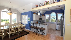 A modern kitchen with white cabinets, a central island with bar stools, and a large window offering a view of the outdoors. - Soundside, Pool, Hot Tub, Community Amenities, MS24, Turtle Whispers (Corolla)