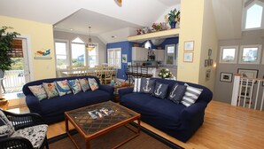 A living room with a blue sofa, a glass coffee table, and a dining area with a white table and chairs. - Soundside, Pool, Hot Tub, Community Amenities, MS24, Turtle Whispers (Corolla)