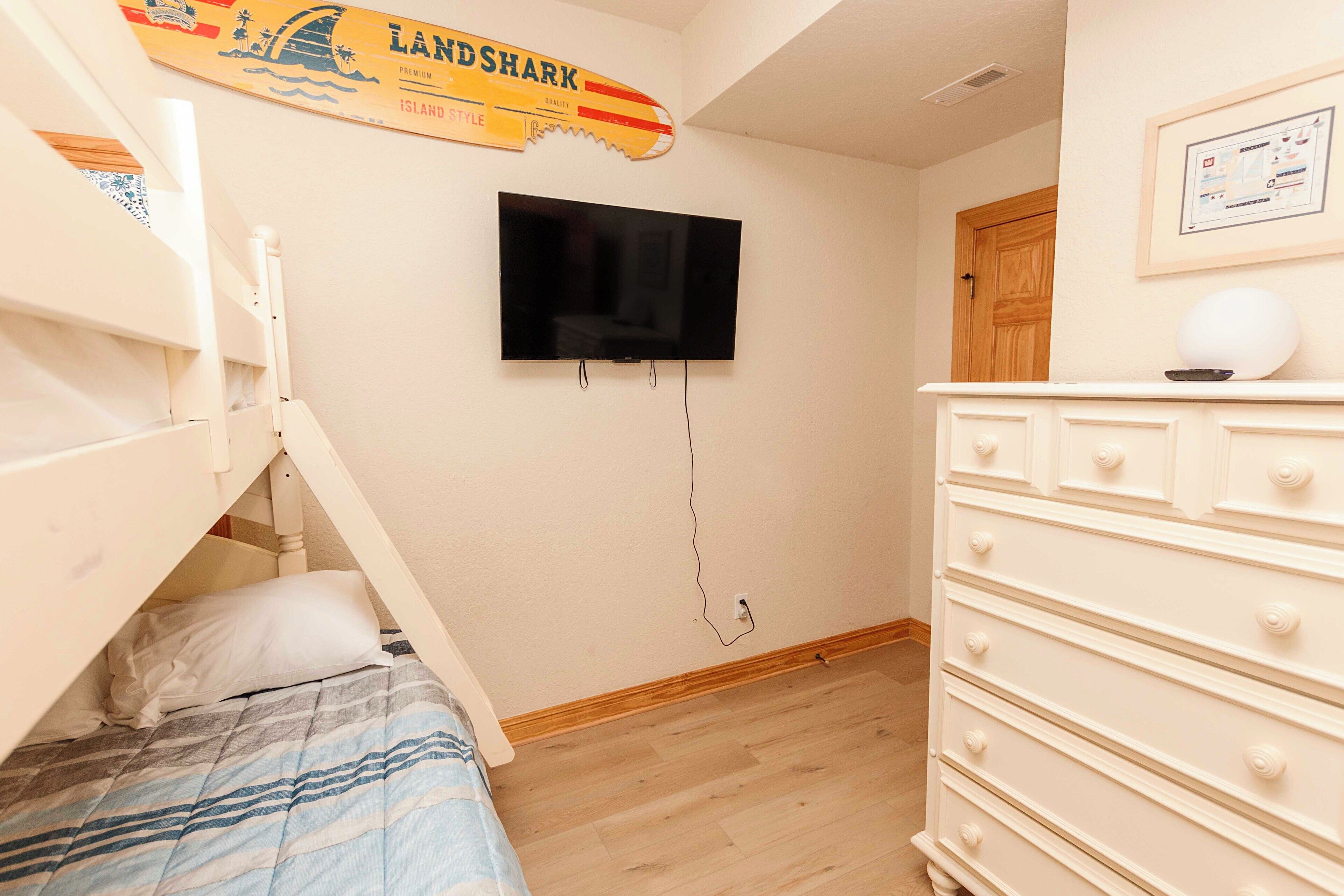 Une chambre avec un lit superposé, un téléviseur à écran plat fixé au mur, une commode et une porte en bois.