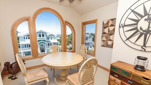 A dining room with a round table and four chairs, large arched windows with a view of houses, and a wooden cabinet with drawers.