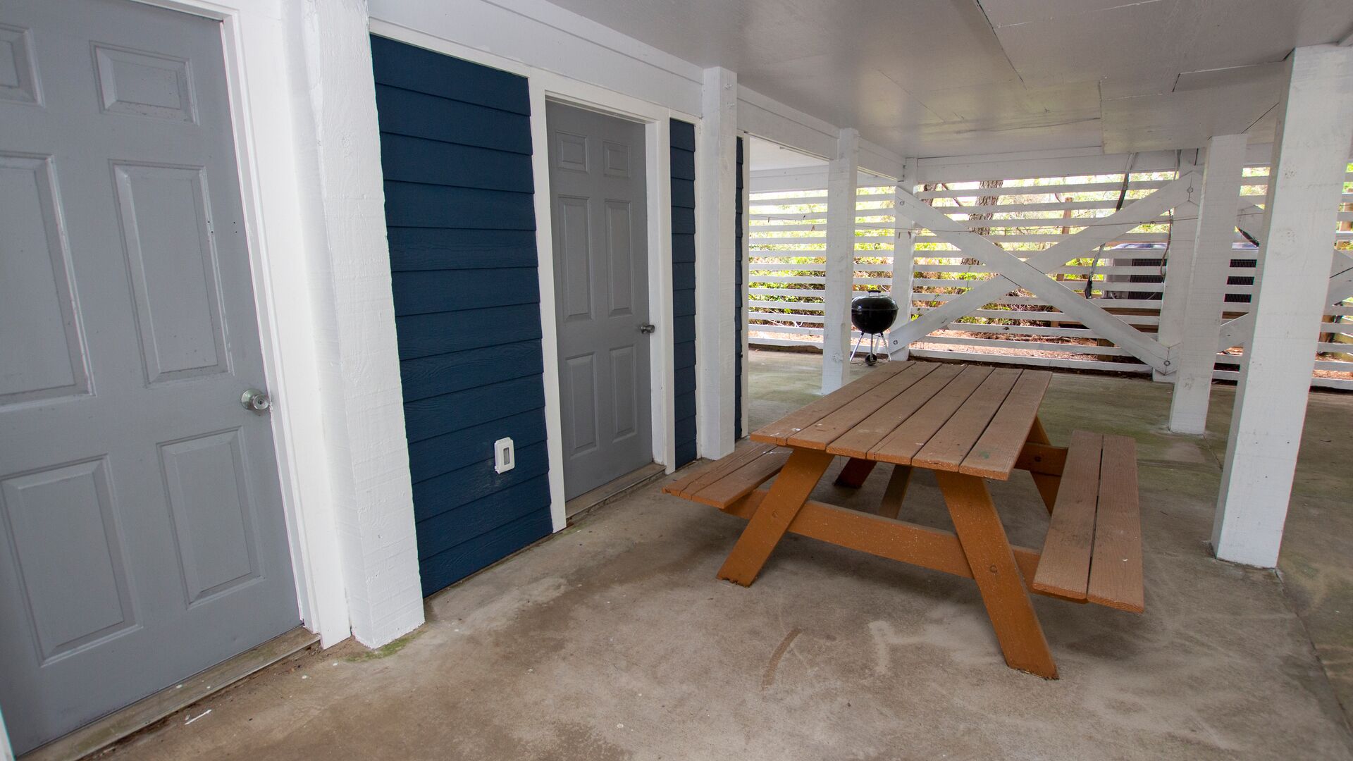 Un área de patio techada con mesa y bancos de madera, una parrilla y una cerca blanca.