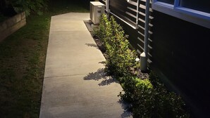 A residential house with a concrete pathway leading to the front door, surrounded by greenery and a wooden fence.