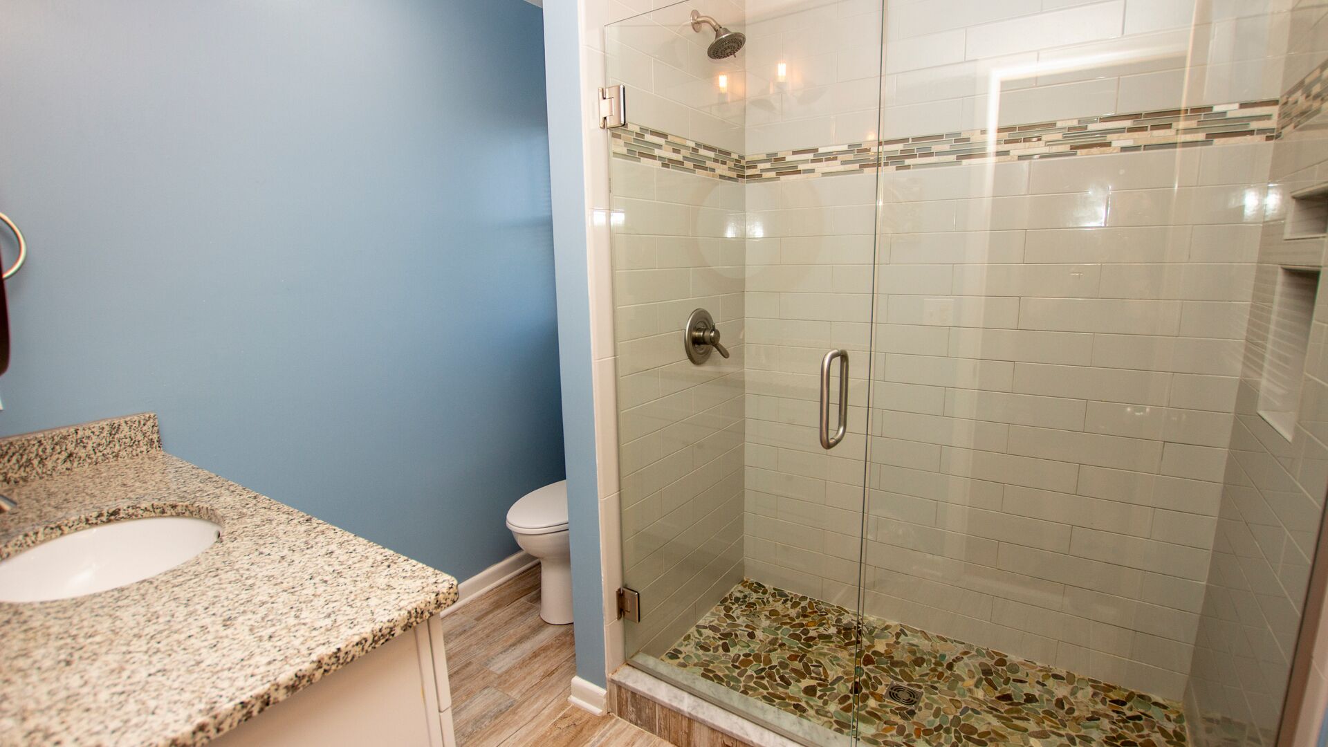 A bathroom with a granite countertop sink, a toilet, and a glass-enclosed shower with mosaic tiles.