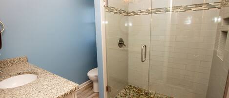 A bathroom with a granite countertop sink, a toilet, and a glass-enclosed shower with mosaic tiles.